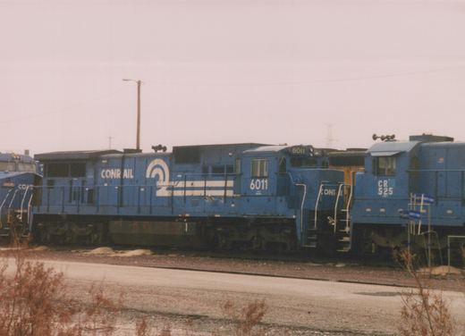 A blue Conrail locomotive numbered 6011 is parked in a rail yard next to other locomotives on a cloudy day.