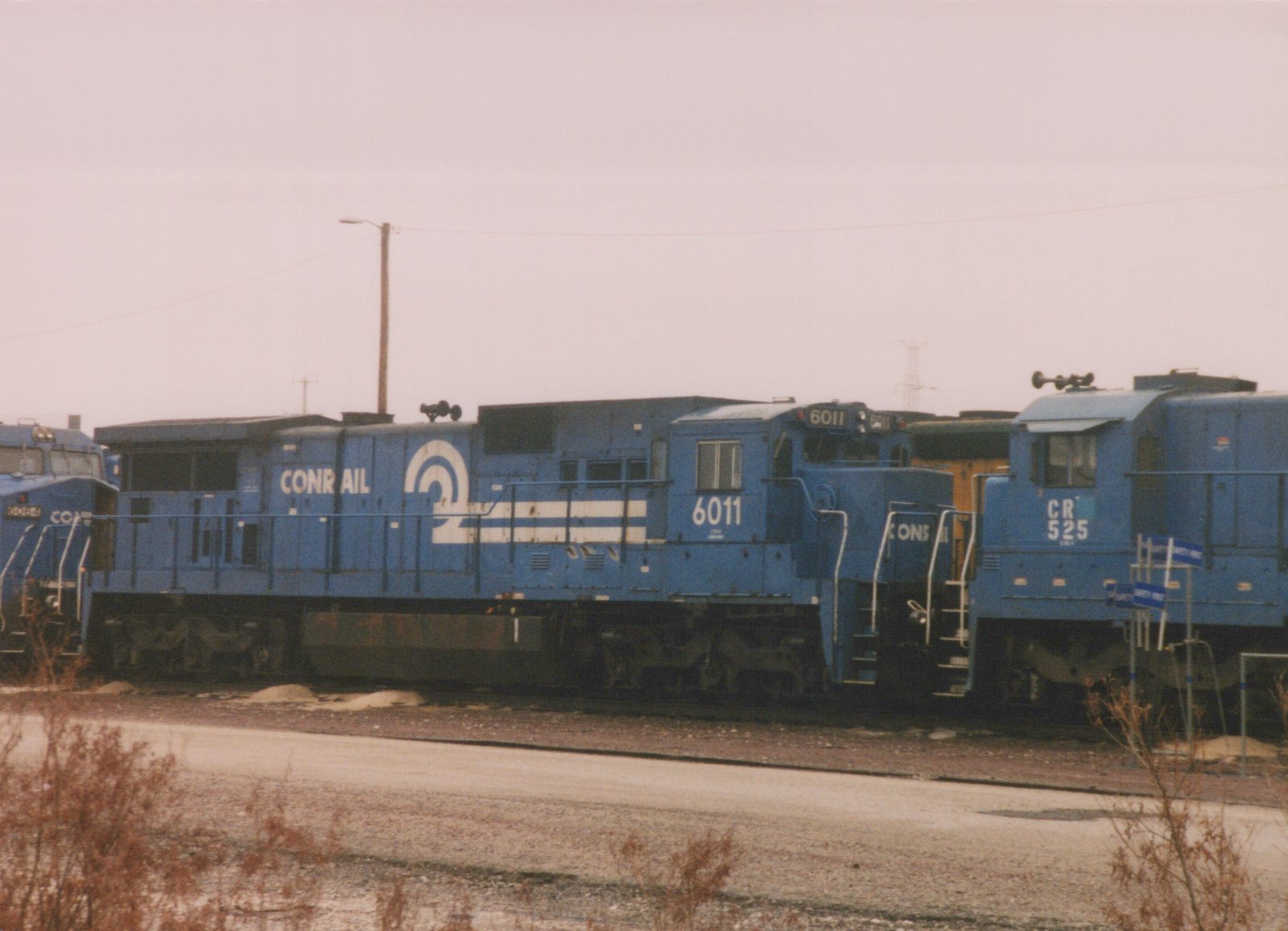 A blue Conrail locomotive numbered 6011 is parked in a rail yard next to other locomotives on a cloudy day.