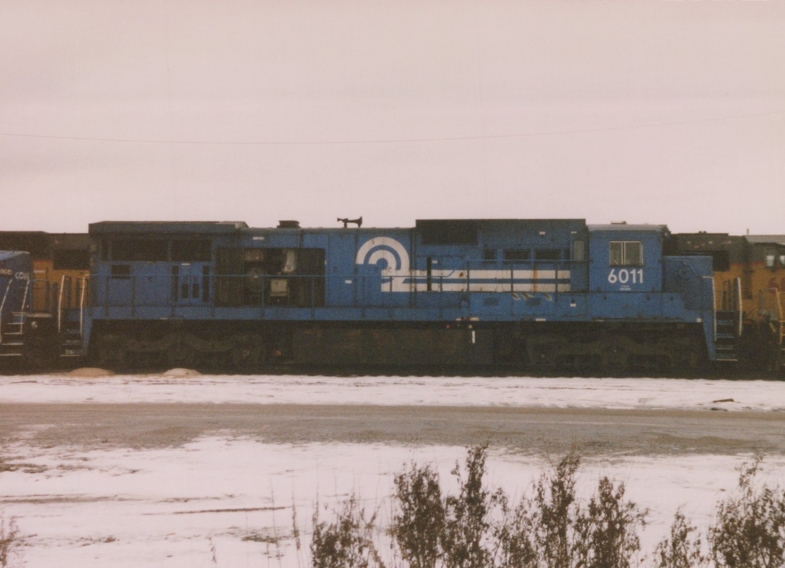A side view of a blue Conrail diesel locomotive, number 6011, standing in a snowy yard with other locomotives partially visible in the background.
