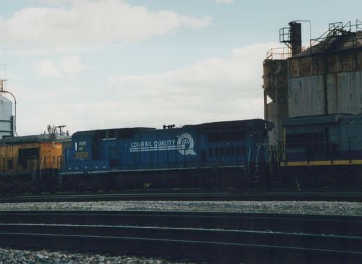 A blue Conrail locomotive with the number 5959 is parked on tracks next to a yellow locomotive in an industrial rail yard under a cloudy sky.