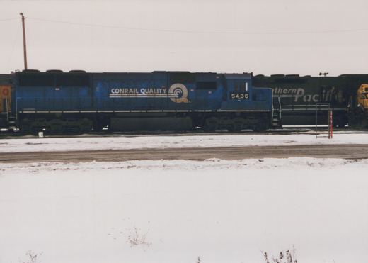 Side view of a blue Conrail diesel locomotive numbered 5436 parked on a snowy railroad yard with another dark green locomotive partially visible behind it.