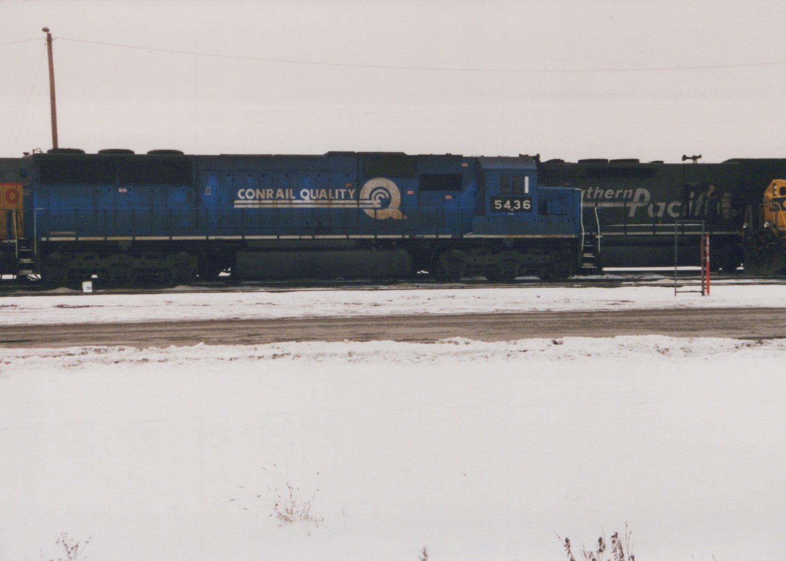 Side view of a blue Conrail diesel locomotive numbered 5436 parked on a snowy railroad yard with another dark green locomotive partially visible behind it.