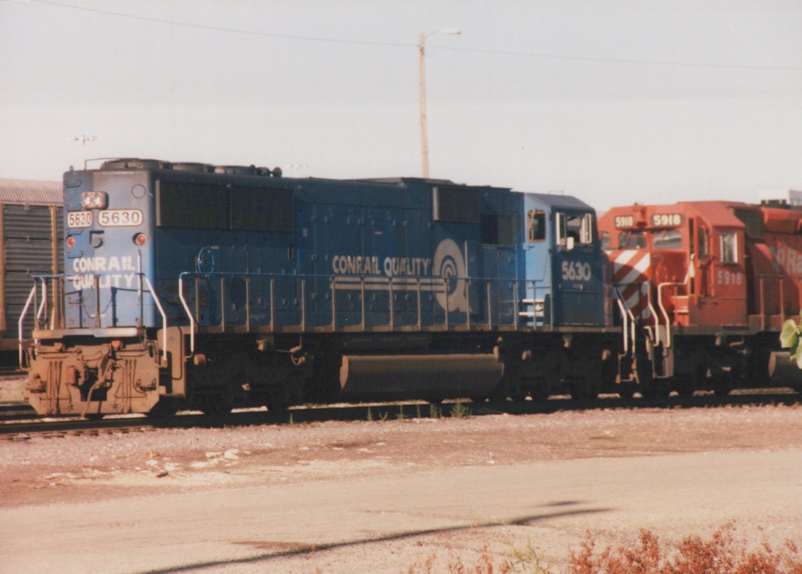 Two Conrail diesel locomotives, one blue and one red, are parked side-by-side in a gravel yard under an overcast sky.