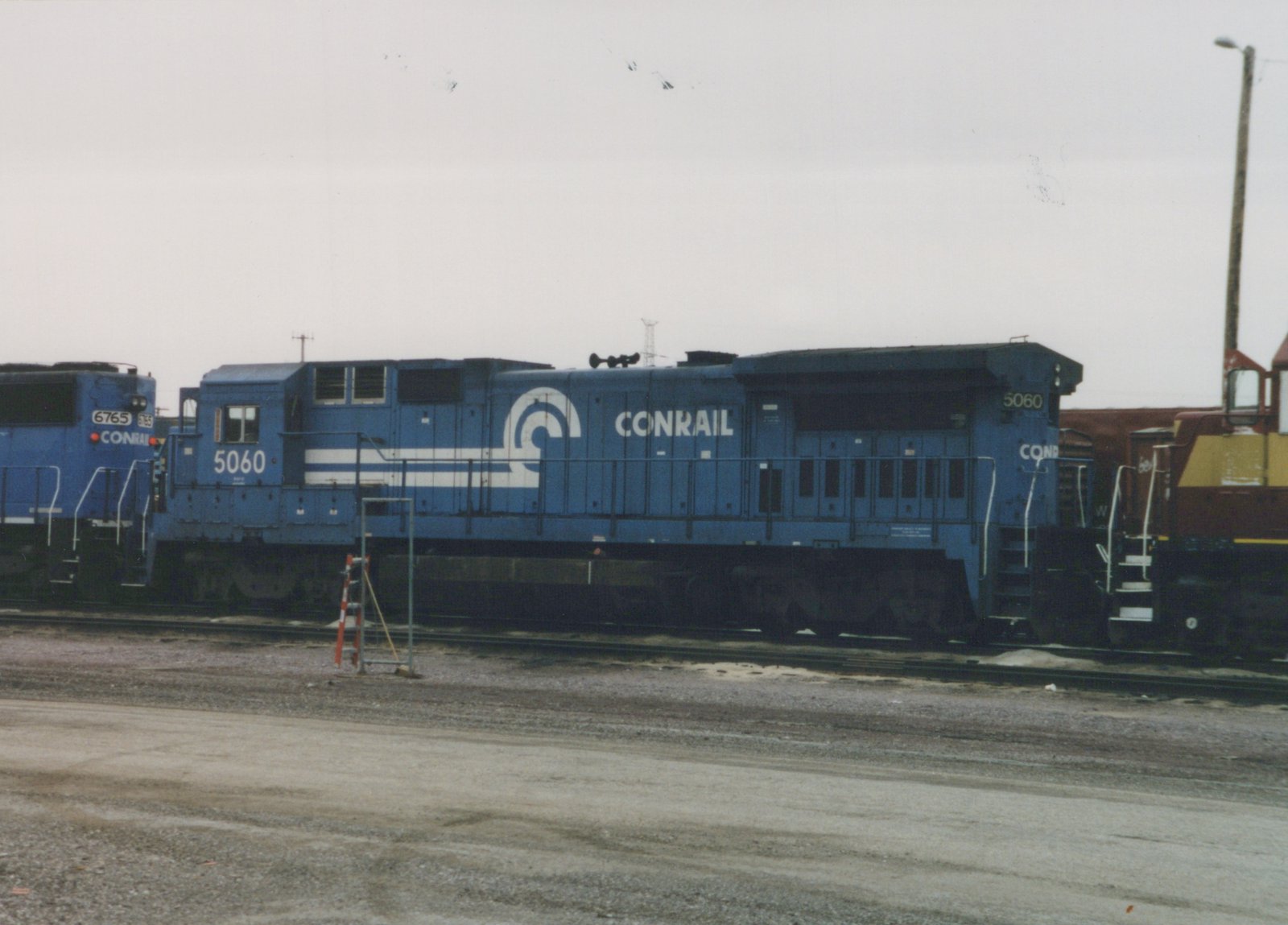Two blue Conrail diesel locomotives with fleet numbers 5060 and 6765 are parked side by side in a gravel rail yard under a gray sky.