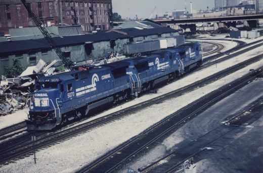 A photograph of three blue Conrail diesel locomotives, numbers 5070 and 5087 visible, moving along tracks in a rail yard with industrial buildings in the background.