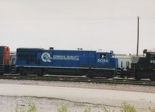 A blue Conrail locomotive with the number 5056 sits on tracks in a rail yard, with other equipment visible nearby.