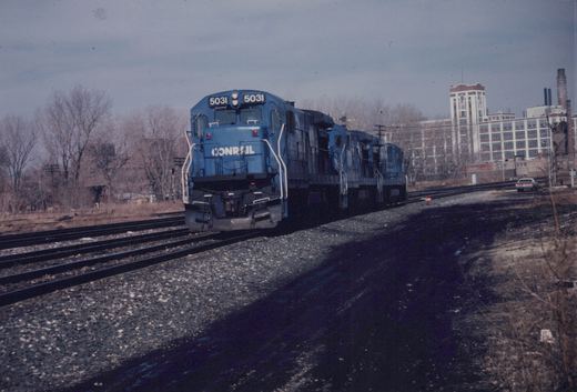 A blue Conrail locomotive numbered 5031 and two matching locomotives are on a set of tracks next to a road with a car, with bare trees and a large brick building in the background under an overcast sky.
