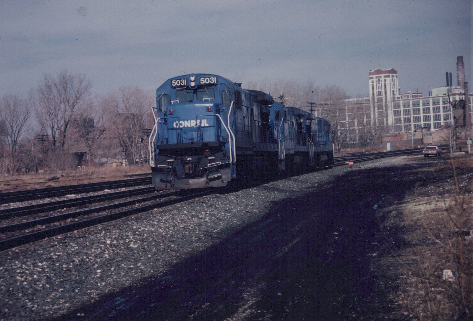 A blue Conrail locomotive numbered 5031 and two matching locomotives are on a set of tracks next to a road with a car, with bare trees and a large brick building in the background under an overcast sky.