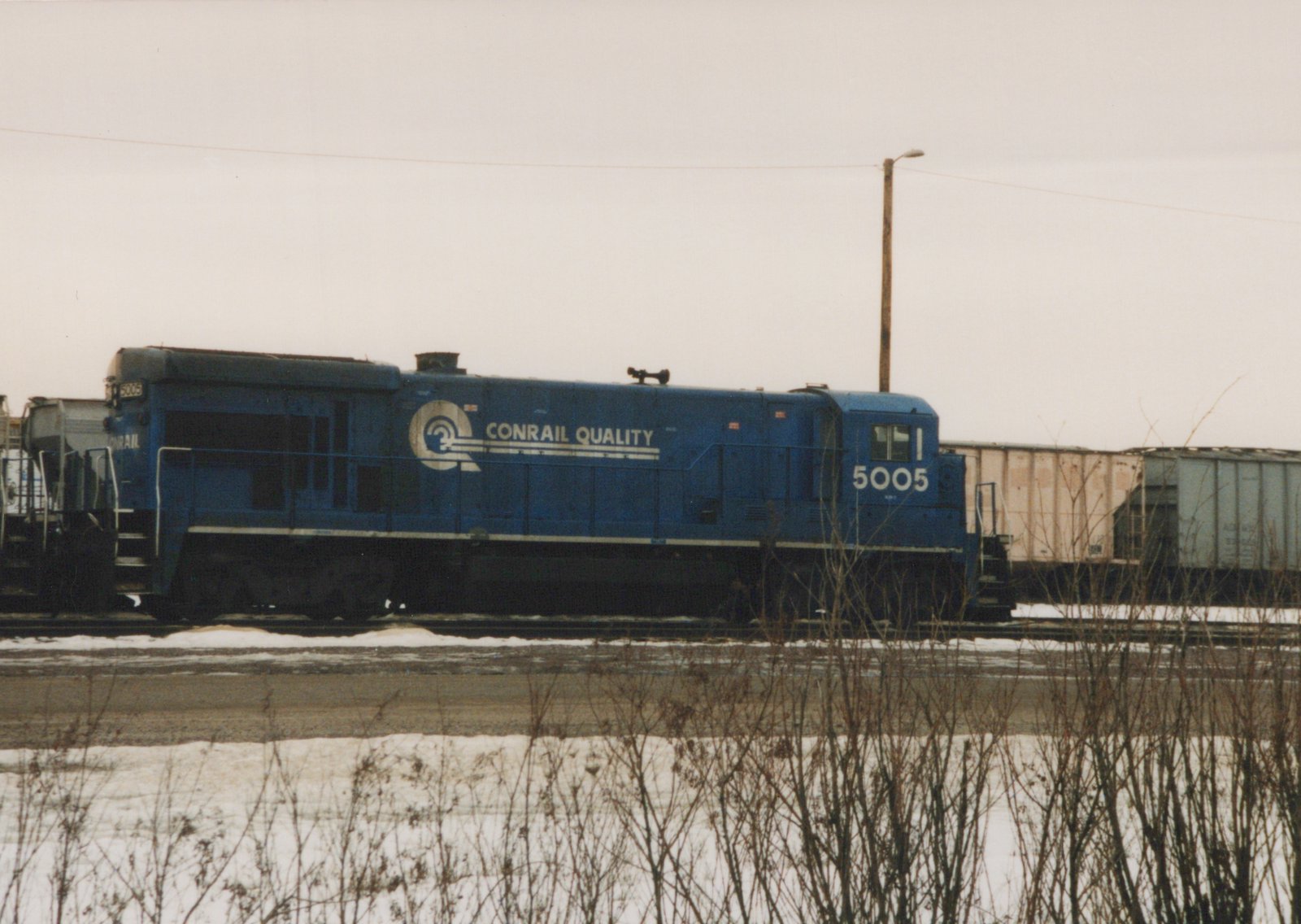 A blue Conrail locomotive numbered 5005 is parked on railroad tracks in a snowy yard, with freight cars visible behind it.