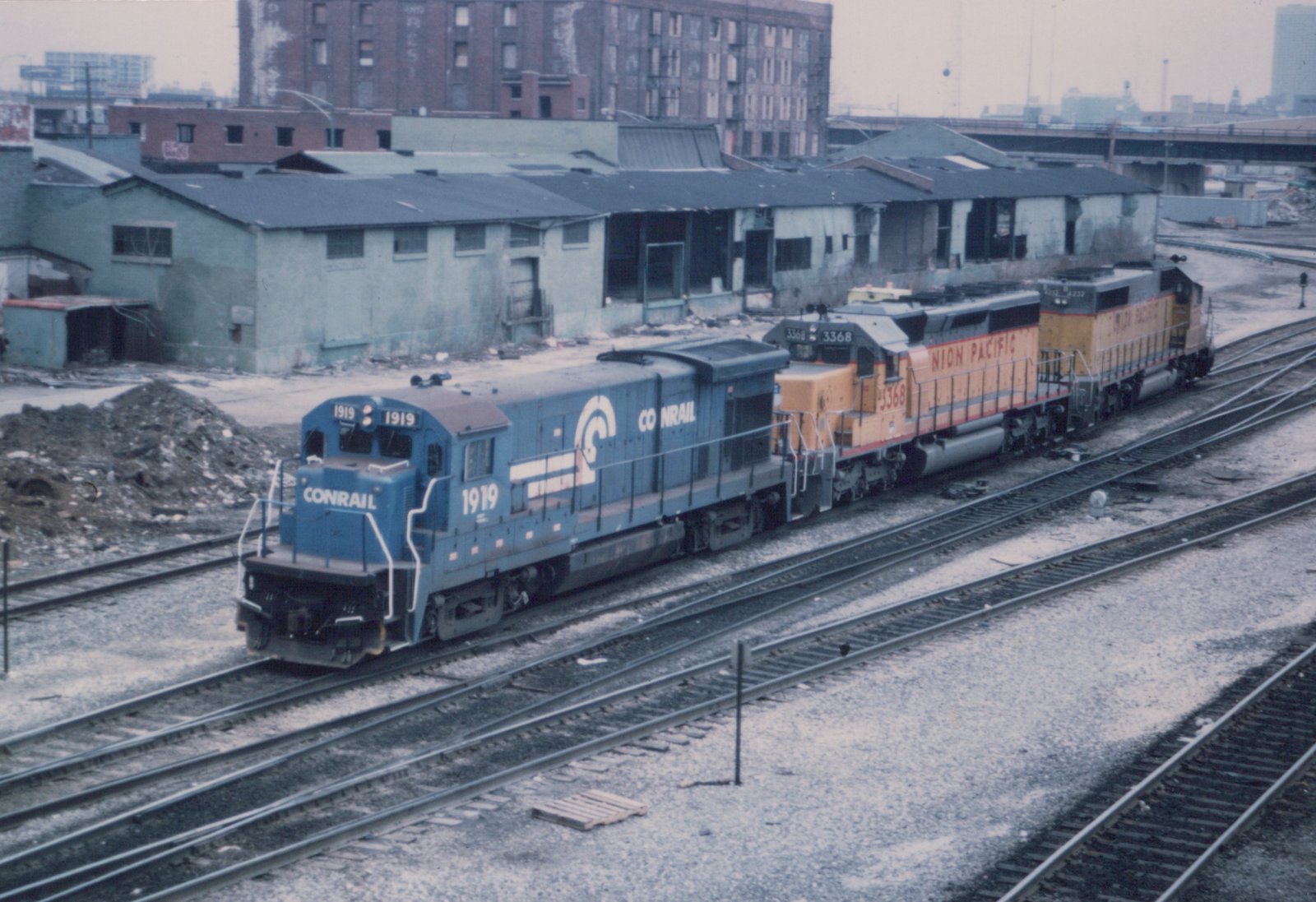 Two diesel locomotives, one blue Conrail and one yellow Union Pacific, on parallel tracks in a snowy industrial yard.