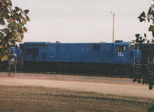 A side view of a blue Conrail diesel locomotive with the number 565, parked in a gravel yard with trees framing the shot.