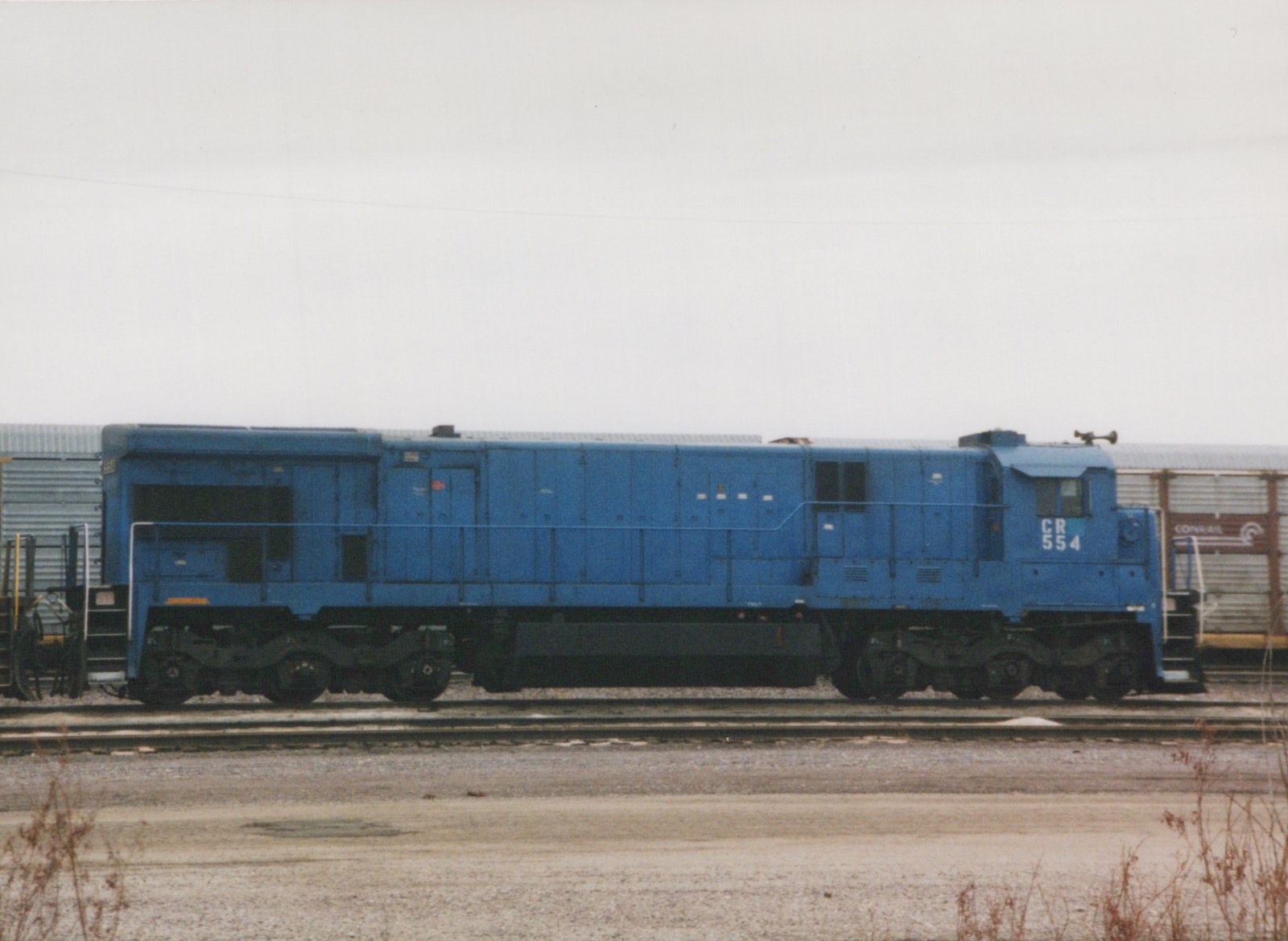 A blue diesel locomotive with the number 554 on its side is parked on tracks in a yard, with a Conrail freight car in the background.