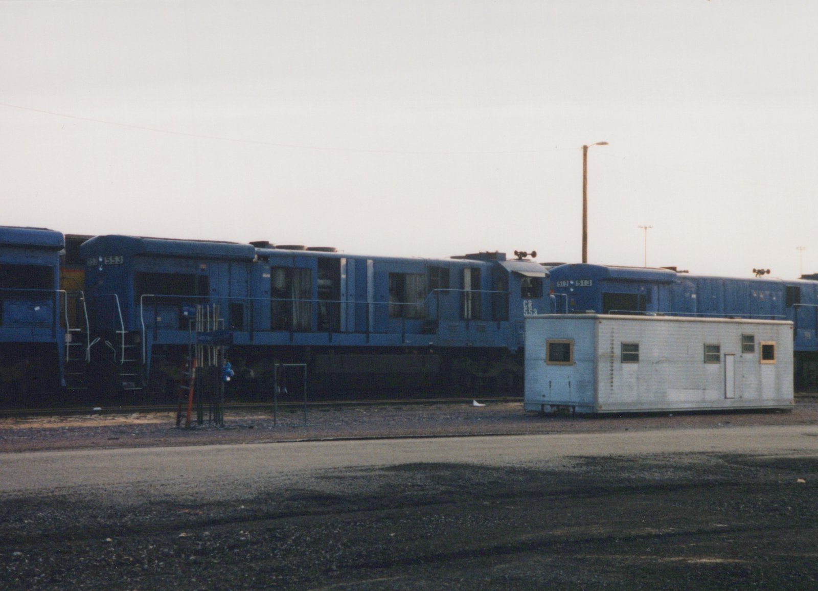 A blue Conrail locomotive numbered 553, likely a GP38-2 model, sits on tracks in a gravel yard with a white trailer nearby.