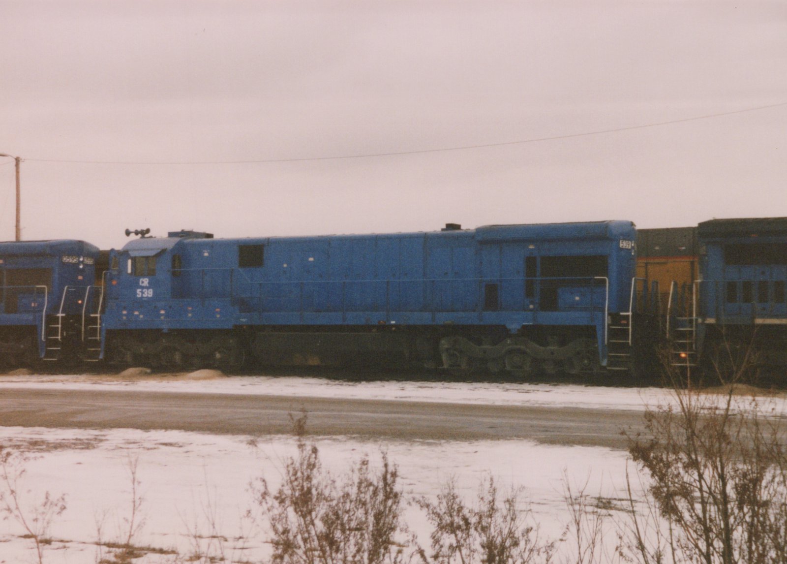 A side view of a blue Conrail diesel locomotive numbered 539 parked in a snow-covered yard with other locomotives nearby under an overcast sky.