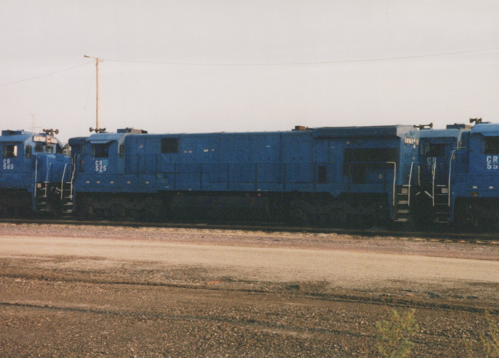 A side view of three blue Conrail diesel locomotives, numbered 565, 525, and 55, parked on tracks in a gravel yard on an overcast day.