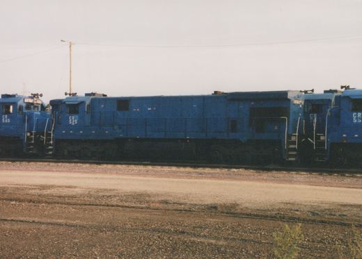 A side view of three blue Conrail diesel locomotives, numbered 565, 525, and 55, parked on tracks in a gravel yard on an overcast day.