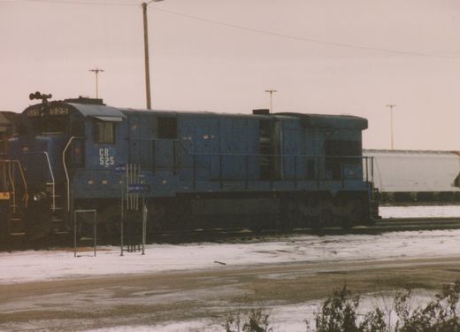 A blue Conrail diesel locomotive numbered 525 is parked on tracks in a snowy yard, with a white freight car visible in the background.