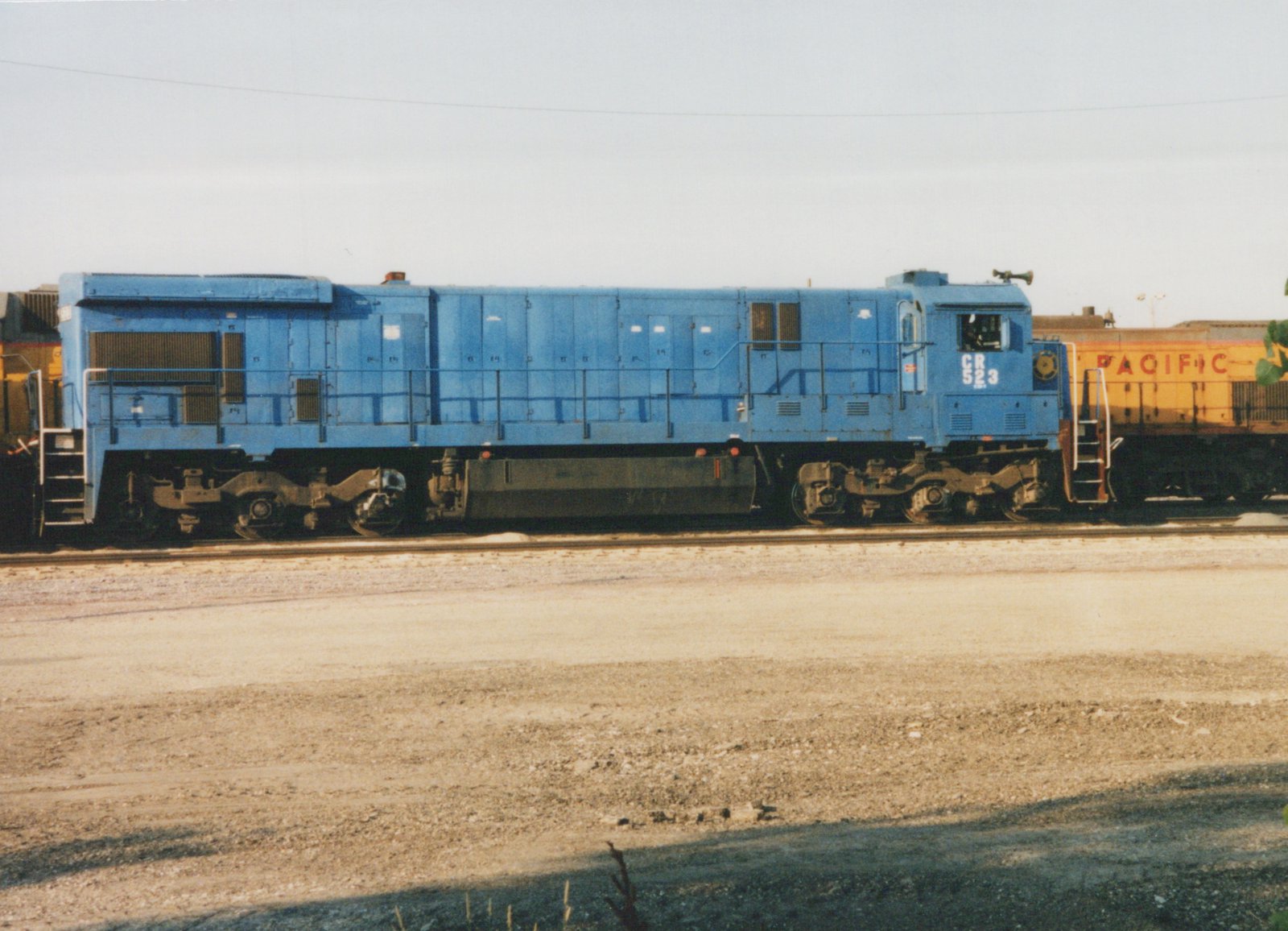 A blue Conrail diesel locomotive, number 523, is parked on a track in a rail yard with a yellow locomotive partially visible in the background.