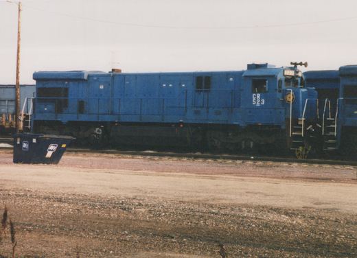 A blue Conrail GP38-2 diesel locomotive numbered 523 is parked in a gravel yard on a cloudy day, with a blue dumpster and freight cars visible in the background.