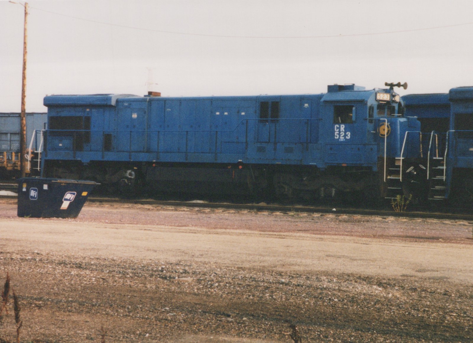 A blue Conrail GP38-2 diesel locomotive numbered 523 is parked in a gravel yard on a cloudy day, with a blue dumpster and freight cars visible in the background.