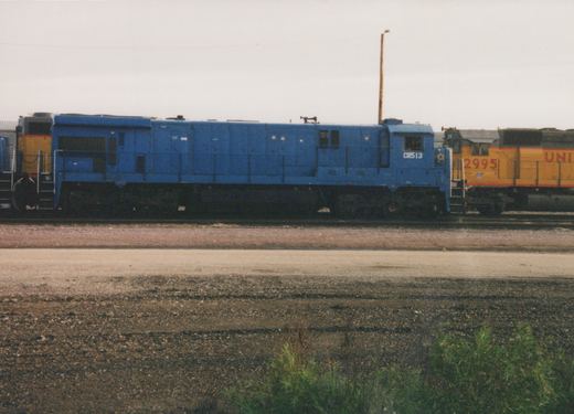 Side view of a blue Conrail diesel locomotive with number 513, parked next to a yellow Union Pacific diesel locomotive with number 2995 in a gravel yard under an overcast sky.