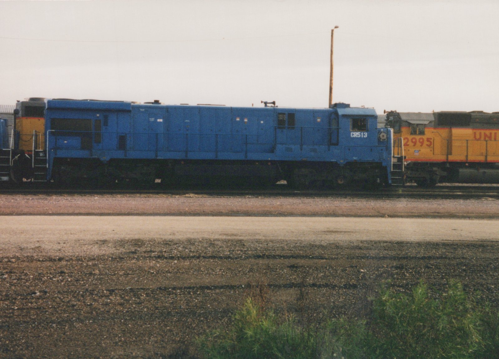 Side view of a blue Conrail diesel locomotive with number 513, parked next to a yellow Union Pacific diesel locomotive with number 2995 in a gravel yard under an overcast sky.