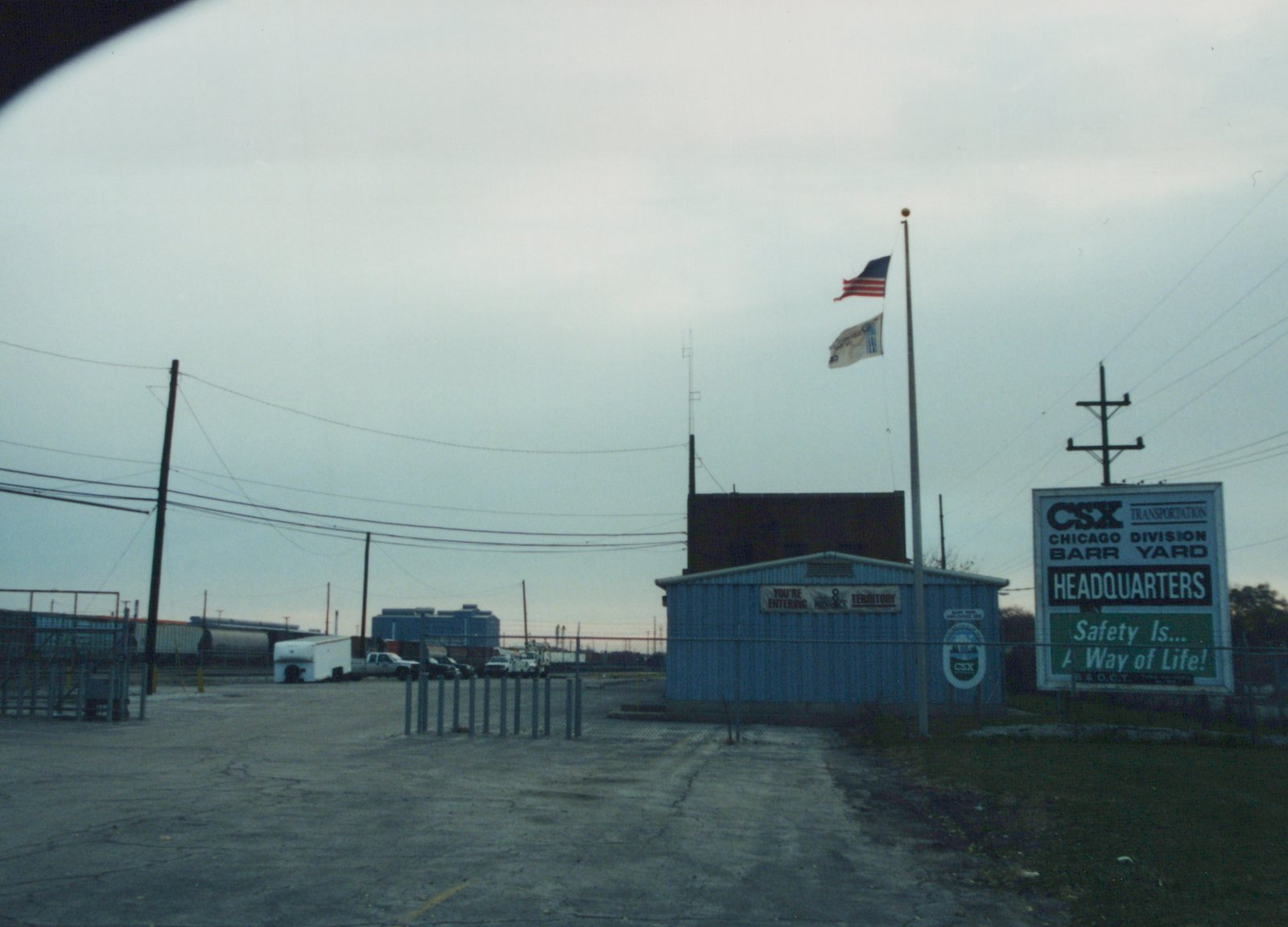 An overcast scene at the CSX Barr Yard headquarters in Chicago, showing a blue building, a large sign, flagpoles, and freight cars in the background.