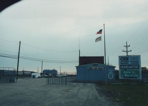 An overcast scene at the CSX Barr Yard headquarters in Chicago, showing a blue building, a large sign, flagpoles, and freight cars in the background.