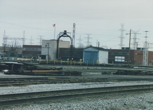 A railroad yard in winter with multiple tracks, an industrial building labeled Acme Steel Company, and a small maintenance vehicle on the tracks under an overcast sky.
