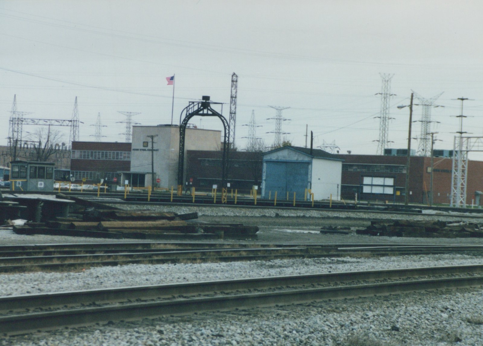 A railroad yard in winter with multiple tracks, an industrial building labeled Acme Steel Company, and a small maintenance vehicle on the tracks under an overcast sky.