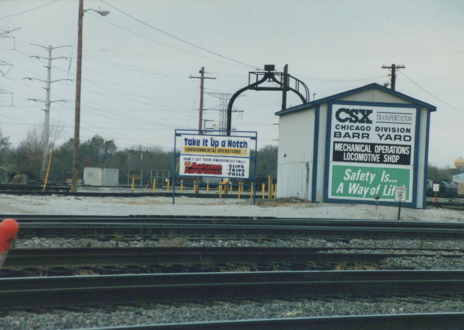 A photograph showing multiple railroad tracks in the foreground and a building in the background with signs for CSX Transportation's Chicago Division Barr Yard and safety messages, under an overcast sky with light snow on the ground.