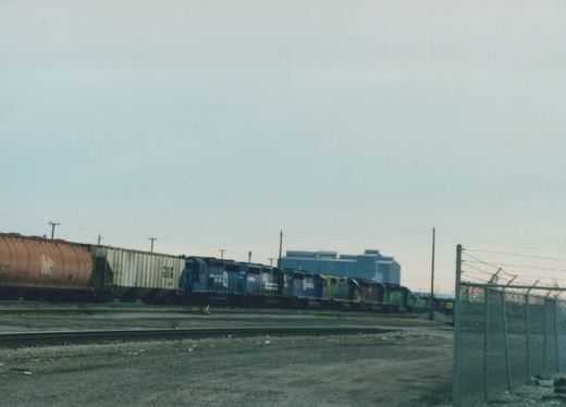 A blue CSX locomotive pulls a freight train including a white boxcar and an orange tank car through a gravel yard next to a chain-link fence under an overcast sky.