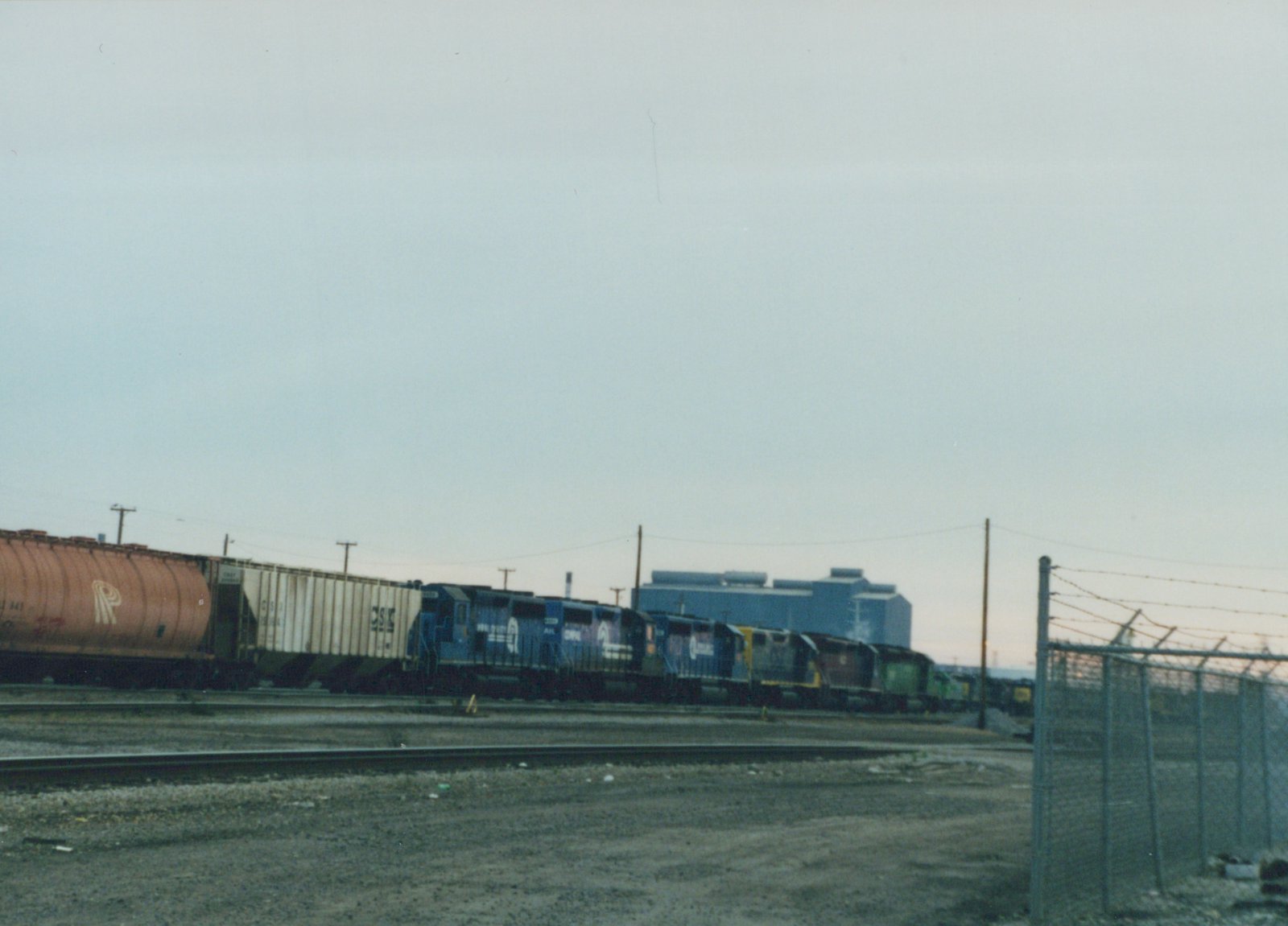 A blue CSX locomotive pulls a freight train including a white boxcar and an orange tank car through a gravel yard next to a chain-link fence under an overcast sky.