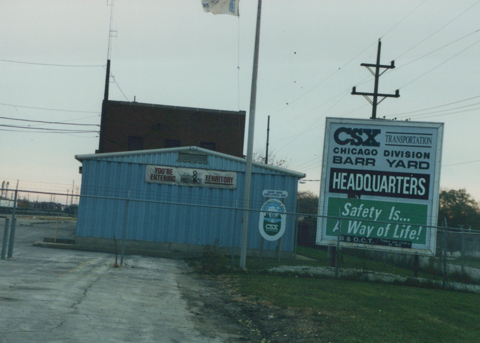 A view of the entrance to CSX's Barr Yard in Chicago, with a large sign and a blue building behind a chain-link fence, under an overcast sky.