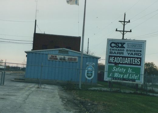 A view of the entrance to CSX's Barr Yard in Chicago, with a large sign and a blue building behind a chain-link fence, under an overcast sky.
