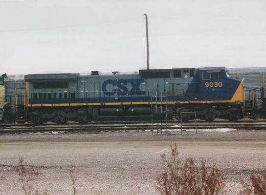 A blue, gray, and yellow CSX locomotive numbered 9030 is parked on a set of tracks in a gravel yard on an overcast winter day.