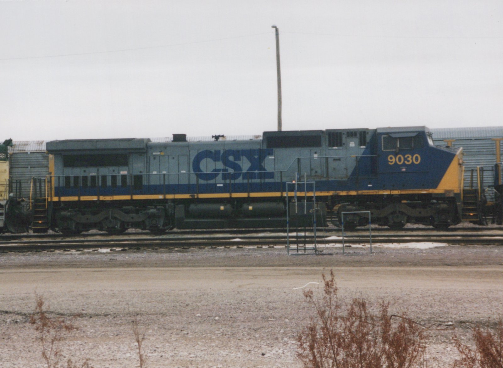 A blue, gray, and yellow CSX locomotive numbered 9030 is parked on a set of tracks in a gravel yard on an overcast winter day.