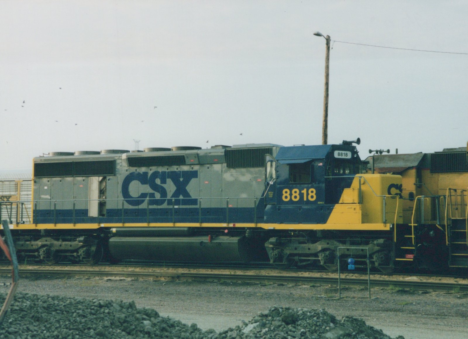 A side view of a CSX Transportation diesel locomotive, number 8818, painted in grey, blue and yellow, sitting on tracks in a gravel yard under an overcast sky.