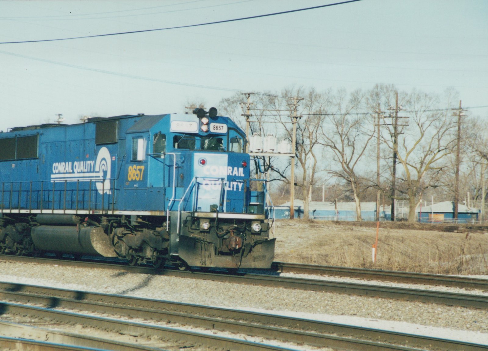 A blue Conrail diesel locomotive number 8657 is moving on a set of train tracks in a winter landscape with bare trees and buildings in the background.