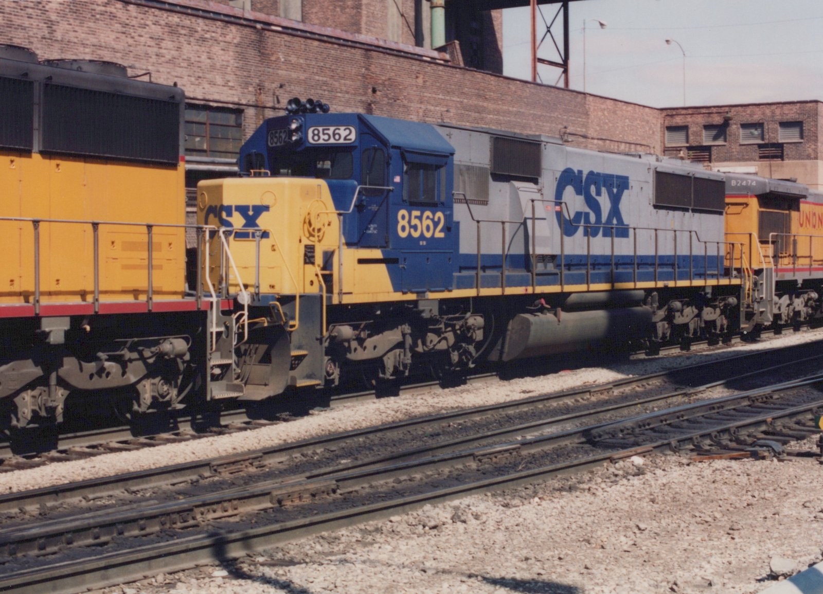Two diesel locomotives, one from CSX and one from Union Pacific, are parked side by side in a railroad yard with brick buildings in the background.