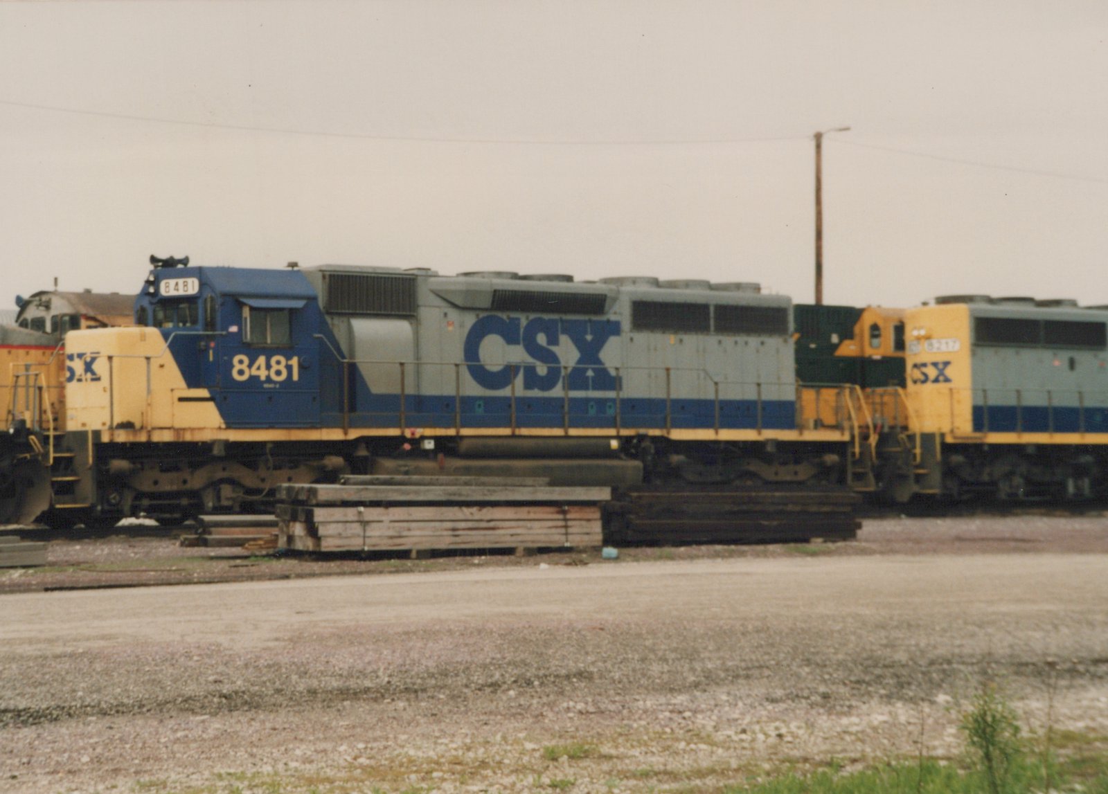 A side view of a CSX locomotive numbered 8481 in a gravel yard, with another CSX engine behind it under an overcast sky.