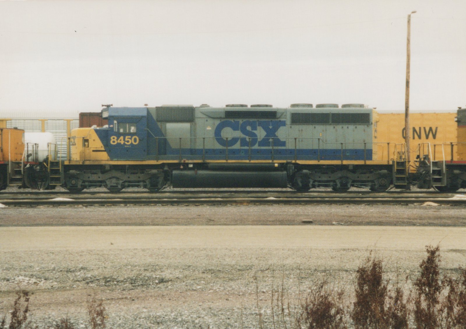 Side view of a blue and yellow CSX diesel locomotive, number 8450, on tracks next to a yellow CNW freight car in a gravel yard under an overcast sky.