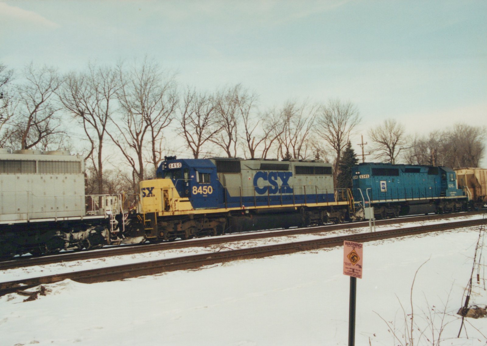 A photo of a CSX freight train with a blue and gray SD40-2 locomotive numbered 8450 and a blue GP38-2 numbered 6345, moving on tracks covered in snow with bare trees in the background.