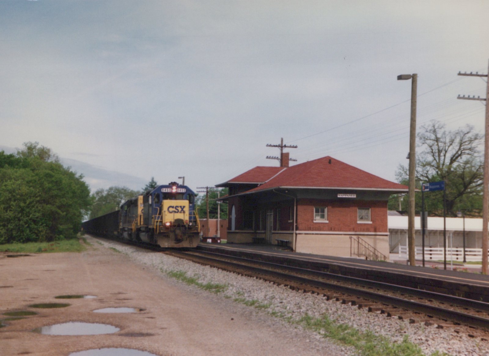 A blue and yellow CSX freight train with locomotive number 8450 passes a brick train station with a red roof in Nappanee, Indiana, on a cloudy day.