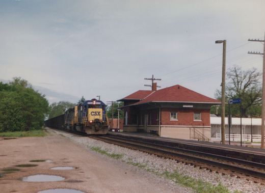 A blue and yellow CSX freight train with locomotive number 8450 passes a brick train station with a red roof in Nappanee, Indiana, on a cloudy day.