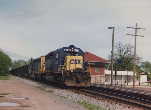 A blue and yellow CSX locomotive numbered 8450 pulls a long line of black freight cars past a small red-roofed station building in Napanee, Indiana, under a cloudy sky.