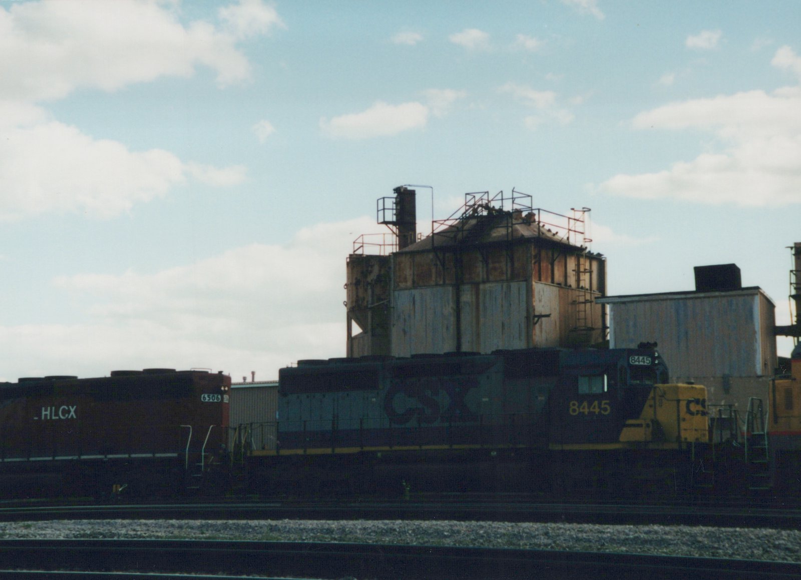 A CSX locomotive numbered 8445 is parked on tracks in a yard, with an HLCX freight car visible behind it and an industrial building in the background under a cloudy sky.