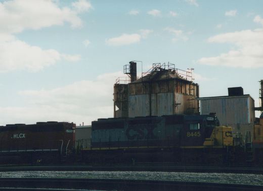 A CSX locomotive numbered 8445 is parked on tracks in a yard, with an HLCX freight car visible behind it and an industrial building in the background under a cloudy sky.