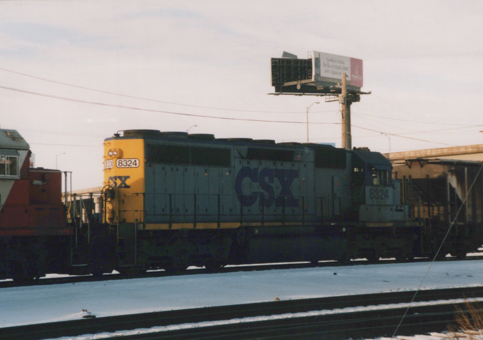 A blue and yellow CSX locomotive numbered 8324 is on a snowy railroad track next to a billboard.
