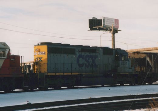 A blue and yellow CSX locomotive numbered 8324 is on a snowy railroad track next to a billboard.
